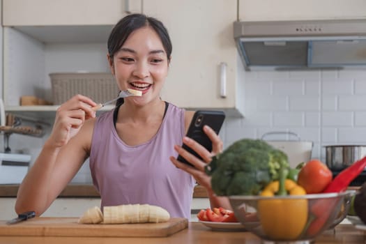 woman with casual clothes making salad and using mobile to count calories and plan diet. Happy woman consulting recipe instructions on smartphone while chopping vegetables in modern kitchen.