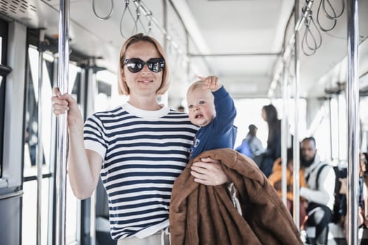 Mother carries her child while standing and holding on to bar holder on bus. Mom holding infant baby boy in her arms while riding in public transportation. Cute toddler traveling with mother