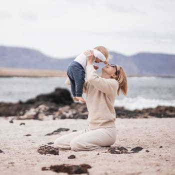 Mother enjoying winter vacations holding, playing and lifting his infant baby boy son high in the air on sandy beach on Lanzarote island, Spain. Family travel and vacations concept.