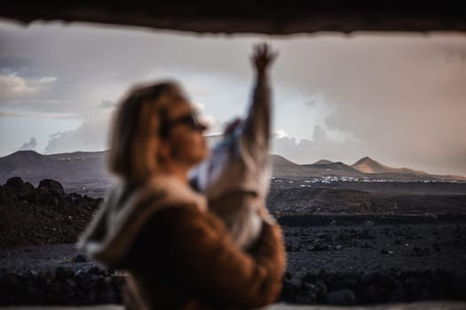 Mother enjoying winter vacations playing with his infant baby boy son on black sandy volcanic beach of Janubio on Lanzarote island, Spain on windy overcast day. Family travel vacations concept