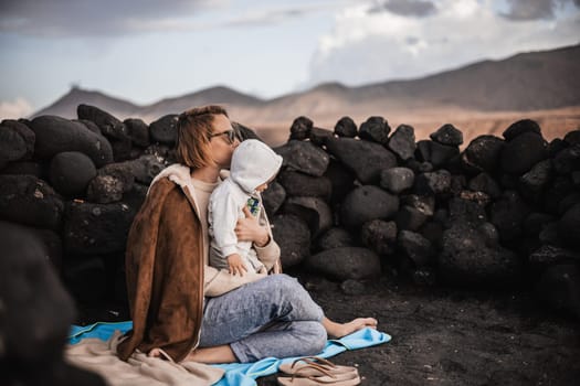 Mother enjoying winter vacations playing with his infant baby boy son on black sandy volcanic beach of Janubio on Lanzarote island, Spain on windy overcast day. Family travel vacations concept