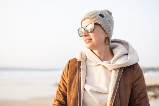 Portrait of young stylish woman wearing brown padded jacket, hoodie, wool cap and sunglasses on long sandy beach in spring