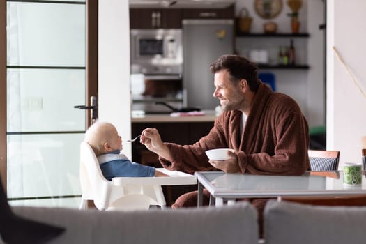 Father wearing bathrope spoon feeding hir infant baby boy child sitting in high chair at the dining table in kitchen at home in the morning