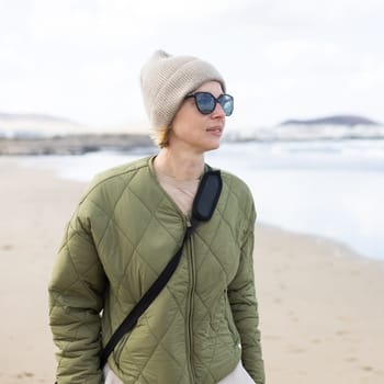 Portrait of young stylish woman wearing green padded jacket, hoodie, wool cap and sunglasses on long sandy beach in spring