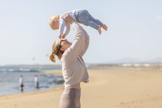 Mother enjoying summer vacations holding, playing and lifting his infant baby boy son high in the air on sandy beach on Lanzarote island, Spain. Family travel and vacations concept