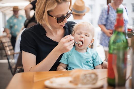 Young caucasian blonde mother spoon feeding her little infant baby boy child outdoors on restaurant or cafe terrace in summer.