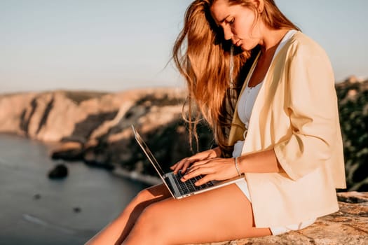 Successful business woman in yellow hat working on laptop by the sea. Pretty lady typing on computer at summer day outdoors. Freelance, travel and holidays concept.