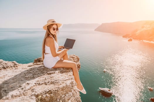 Successful business woman in yellow hat working on laptop by the sea. Pretty lady typing on computer at summer day outdoors. Freelance, travel and holidays concept.