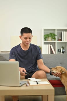 Smiling asian man in casual clothes playing with lovely dog while sitting on sofa living room.
