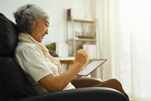 Beautiful middle age woman using digital tablet on comfortable armchair. People and technology concept.