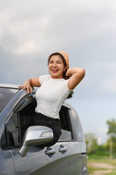 Cheerful young woman leaning out from car window enjoying road trip on a summer day.