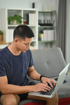 Image of male freelancer in casual clothes working on laptop while sitting on sofa at home.