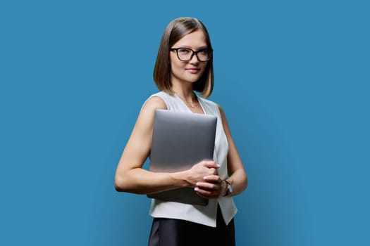 Young confident woman with laptop computer in hands on blue studio background. Smiling female with glasses looking at camera. Using mobile application for work business study leisure technology people