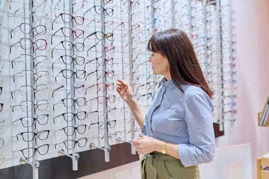 Middle-aged woman in optician's store buying, choosing glasses frames, near display case with vision eyeglasses spectacles. Optics, ophthalmology, age vision problems