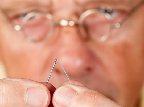 Macro image of a needle and cotton thread as senior man with glasses tries to thread the cotton