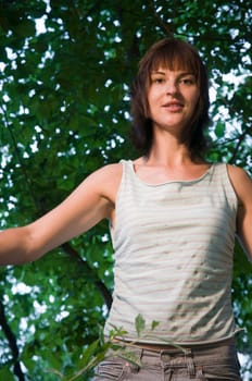 Upper body shot of a young brunette woman with medium length hair and sleeveless top and green tree leaves in the background