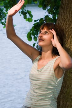 Upper body portrait of a young Caucasian woman looking up in shock,her hands stretched upwards leaning against a tree at a lake in Potsdam (Griebnitzsee)