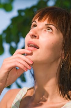 Head-and-shoulders portrait of a young,Caucasian,questioningly upward-looking,amazed woman in side view with her hand on her chin and open mouth standing under trees at a lake in Potsdam (Griebnitzsee)