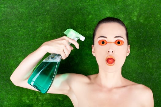 Closeup of a woman wearing tanning bed glasses with strawberry in mouth and spray bottle in hand against green