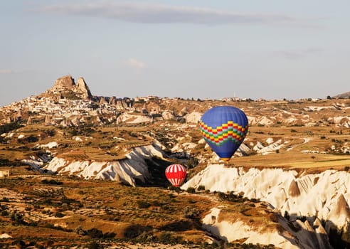 Uchisar backdrop red and white plus blue multi colored hot air balloons navigate a gorge during low level flight, blue sky, wilderness, desert plants, limestone cleft, volcanic region, landscape, crop space and copy area