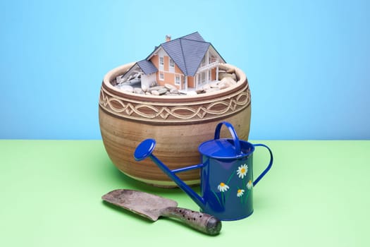 Gardener still-life representing dream about own house - can, small garden shovel and model of the house in flowerpot.
