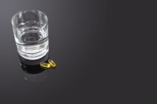 A couple of yellow vitamin pills on a black glass table with a glass of water at the side