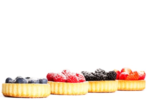 Sugared blueberries on tartlet cake in front of other berries on white background