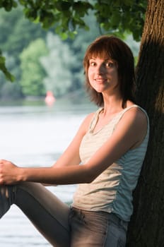 Upper body shot of a young woman leaning against a tree at a lake (Griebnitzsee in Potsdam / Germany) at sunset looking at the camera