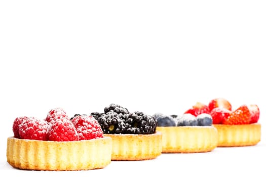 Sugared raspberries on tartlet cake in front of other berries on white background