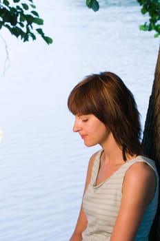 Side torso shot of a young woman in a short top by a lake and lowered thoughtful gaze,leaning against a tree trunk