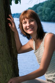 Upper body shot of a young woman leaning against a tree at a lake (Griebnitzsee in Potsdam / Germany) at sunset looking at the camera