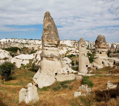 square format group of limestone fairy chimney homes near Goreme Cappadocia Turkey, volcanic lava hills in the background, cloudy blue sky with crop space and copy space