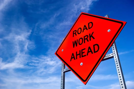 Image of a bright orange road work ahead sign against a blue sky with light clouds
