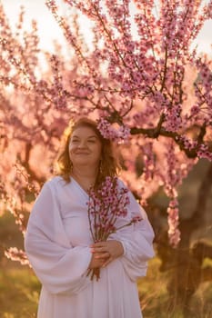 Woman blooming peach orchard. Against the backdrop of a picturesque peach orchard, a woman in a long white dress enjoys a peaceful walk in the park, surrounded by the beauty of nature