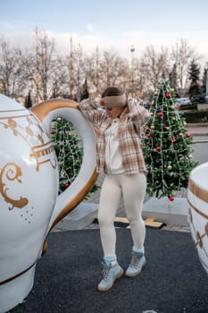 Woman Christmas Square. She stands near a large white cup, dressed in a light suit. With trees decorated with Christmas tinsel in the background.