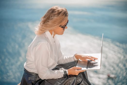 Business woman on nature in white shirt and black skirt. She works with an iPad in the open air with a beautiful view of the sea. The concept of remote work