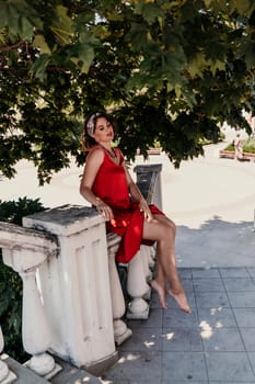 woman in a red silk dress and a bandage on her head smiles against the background of the leaves of a tree. She is leaning on the coop and looking into the camera. Vertical photo