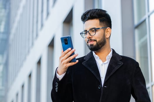 Handsome Indian businessman using smartphone typing text answering messages chatting online looking mobile screen app outdoors. Arabian Hindu freelancer guy in downtown city street. Business people