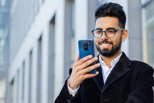 Stylish Indian businessman using smartphone typing text answering messages chatting online looking mobile screen app outdoors. Arabian Hindu freelancer guy in downtown city street. Business people