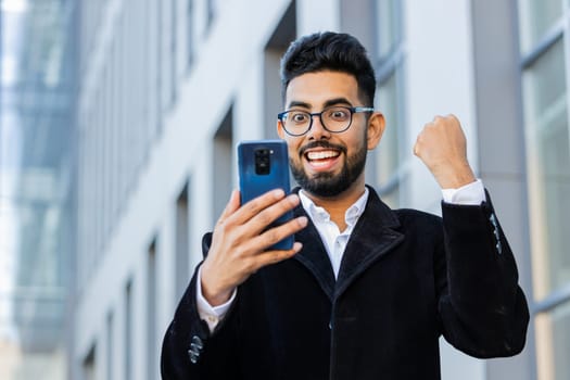 Happy Indian businessman use mobile smartphone celebrating win good message news, lottery jackpot victory, online outdoors. Arabian Hindu freelancer in downtown city street. Business people lifestyle