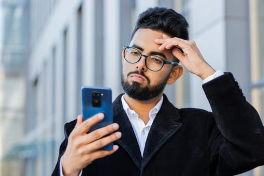 Indian businessman use smartphone typing browsing, loses surprised by bad news, fortune loss, fail bankruptcy outdoors. Arabian Hindu freelancer guy in downtown city street. Business people lifestyle