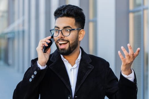 Phone call, good news, gossip. Happy surprised Indian businessman in pleasant conversation on smartphone, enjoying talking outdoor. Arabian Hindu guy in downtown city street. Business people lifestyle