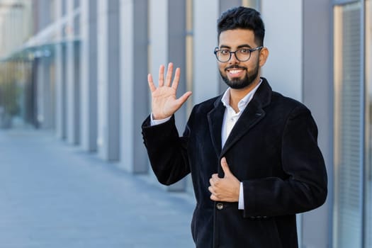 Hello. Indian businessman smiling friendly at camera, waving hands gesturing hi, greeting or goodbye welcoming with hospitable expression outdoors. Arabian Hindu freelancer guy in downtown city street
