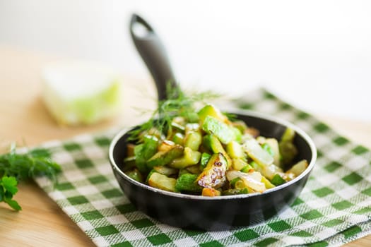 Sliced zucchini fried with onions and herbs in a frying pan on a wooden table.