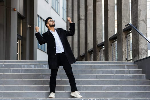 Happy Indian businessman runs down the stairs celebrating success, winning, career achievement, good news, luck, victory, successful signing of contract. Arabian Hindu freelancer guy in downtown city