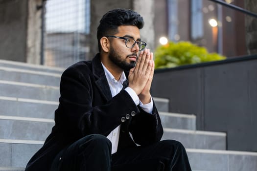 Indian businessman clasping hands wishing luck praying to God asking blessing help forgiveness wish luck outdoor. Arabian Hindu freelancer guy in downtown city street. Business people lifestyle