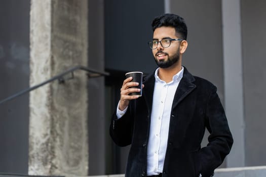 Happy bearded Indian businessman enjoying morning coffee hot drink and smiling outdoors. Relaxing, taking a break. Arabian Hindu freelancer guy drinking coffee to go in downtown city street. Lifestyle