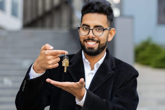 Indian businessman real estate agent lifting hand showing keys of new home house apartment buying or renting property, mortgage loan, investment outdoors. Arabian Hindu freelancer guy in downtown city