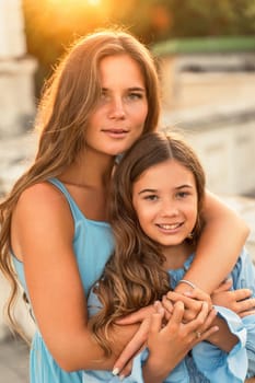 Portrait of mother and daughter in blue dresses with flowing long hair against the backdrop of sunset. The woman hugs and presses the girl to her. They are looking at the camera