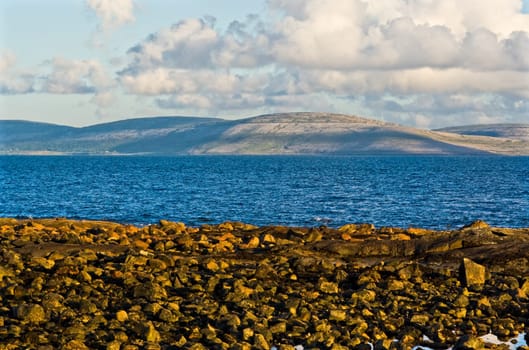 Galway Bay in Ireland from the town of Spiddal with The Burren across the bay. Photo is layered from front to back with Rocks, Galway Bay, The Burren, and sky.